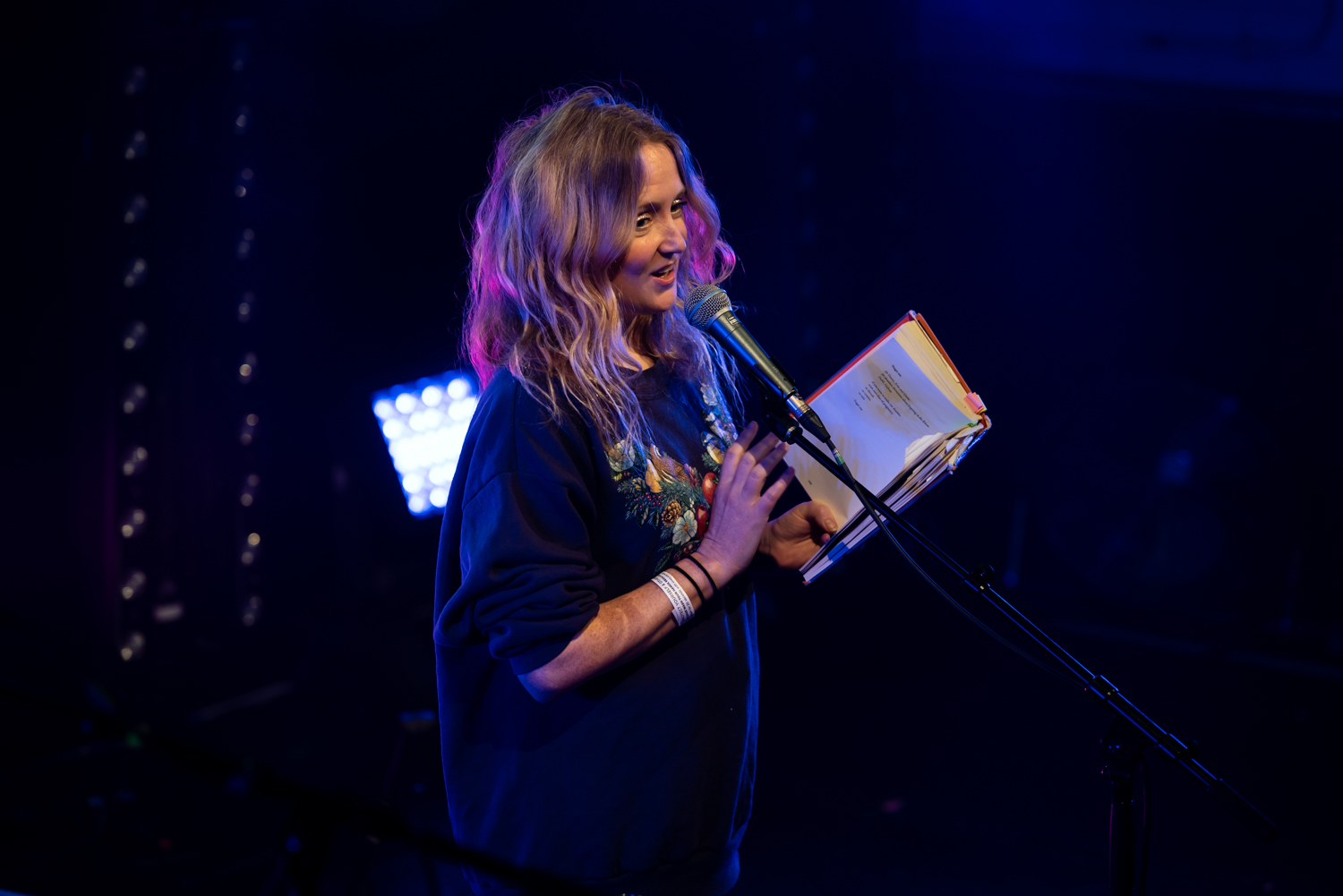 Photo of Hollie on stage wearing a lush purple Christmassy jumper with a sort of fruit bouquet on holding her book and drenched in a purply pink stage spotlight as she reads smiling into the microphone